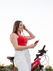 Fototapeta na wymiar Teenage girl standing next to her bike listening to the music in headphones in the park at sunset