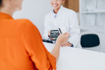 Young female practitioner or nurse with face protective mask working at clinic reception desk. She is helping to patient to make contactless payment for medical services.