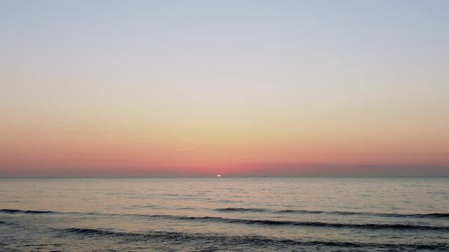 夕日が沈む水平線を眺めながら浜辺に恋人同士が座っている。穏やかな青空のもと白波と赤く染まる夕陽が美しい。