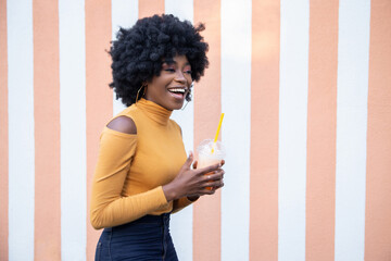 Smiling African American woman with curly hair, holding is orange cocktail in a plastic cup with yellow straw and looking away, leads healthy lifestyle, posing on striped background.