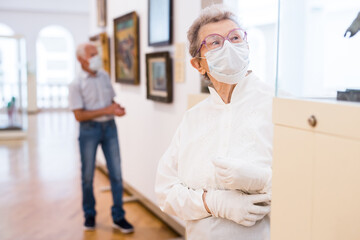 elderly European woman  in mask protecting against covid examines paintings on display in hall of art museum