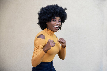 Positive pretty African American woman with curly hair, raises clenched fists, ready to give punch...