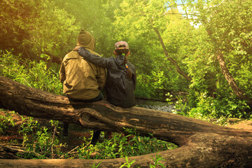 Fototapeta premium A man and a girl sit on a fallen tree and admire the beautiful forest stream. Outdoor activities and hiking in the woods with children. Daughter hugs father. Family adventures in the summer holidays.
