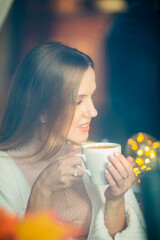 Beautiful young woman is drinking coffee. Attractive girl in a cozy cafe or at home. Pretty woman with a cup of cappuccino in her hands. Close-up portrait. Soft focus and shallow depth of field.