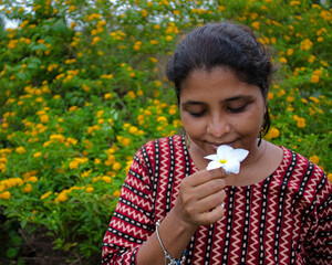 Pretty brunette smells a white flower wearing a designer top and bracelets, with yellow flowers in background during springtime
