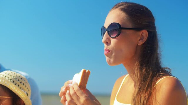 family, leisure and people concept - happy mother, father and two daughters having picnic on summer beach and eating sandwiches