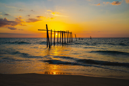 The view of the old wooden bridge decayed in the middle of the ocean in the evening. The sun is setting, making the sky a golden yellow. The waves hit the shore with orange light. There is copy space.