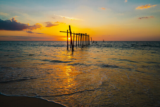 The view of the old wooden bridge decayed in the middle of the ocean in the evening. The sun is setting, making the sky a golden yellow. The waves hit the shore with orange light. There is copy space.