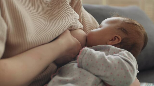 Close up of unrecognizable woman sitting on couch at home and breastfeeding her mixed-race infant daughter lying on her knees