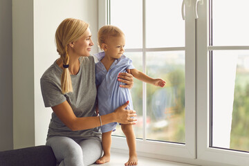 Happy young mother and little toddler boy observing neighborhood from windowsill