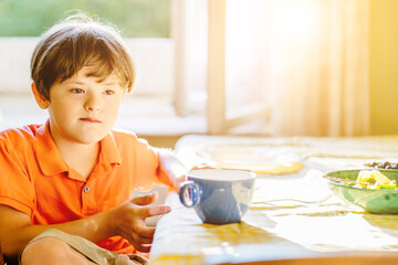 Thoughtful child boy wearing orange t-shirt with down syndrome sitting at table at home.