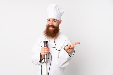 Redhead man using hand blender over isolated white background pointing finger to the side