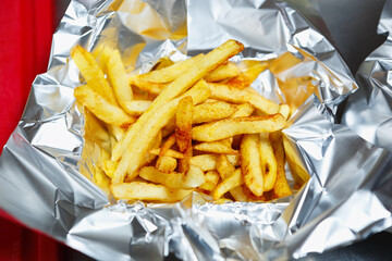 Delicious French fries served in tin foil for fast food delivery.Delicious golden potato chips cooked in deep fryer oven for take away dinner meal.Fat junk foods with big amount of calories