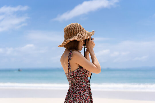 A young Asian woman wearing a floral dress and a straw hat poses for a photograph by the film camera at the seaside in the daytime.
