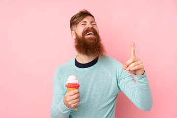 Redhead man with long beard holding a cornet ice cream over isolated pink background intending to...