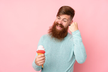 Redhead man with long beard holding a cornet ice cream over isolated pink background celebrating a...