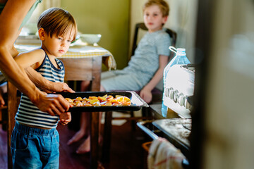 Cute little preschooler boy in striped t-shirt help his mother at kitchen. Cook at home. Young woman pouring oil at a baking tray before oven. Her son helping hold. Family cooking dinner together