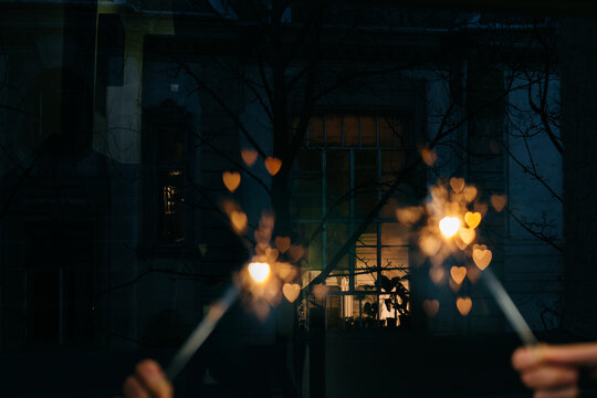 Two hands holding two sparklers in the window making heart shape bokeh in front of a building
