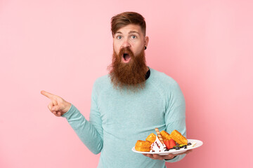 Redhead man with long beard holding waffles over isolated pink background surprised and pointing side