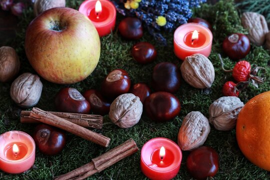 Colorful esoteric autumn fall background filled with September harvest and nature finds. Apple, orange, cinnamon sticks, walnuts, chestnuts and orange colored burning candles on green moss background
