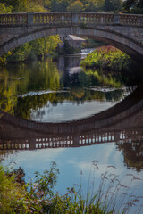 Fototapeta premium Stone Bridge Reflected in the White Cart River in Glasgow With Watermill in the Background on a Sunny Day in Autumn