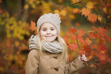 Happy little girl holding a maple tree branch in the autumn park. Pretty blond girl wearing beige coat, knitted cap and scarf smiling.