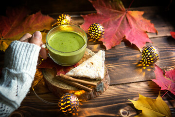 Green Japanese matcha tea with foam in transparent Cup on wooden table in autumn still life. Women's hand with long white sweater sleeves are warmed by drink. Focus on Cup. Warm atmosphere and comfort