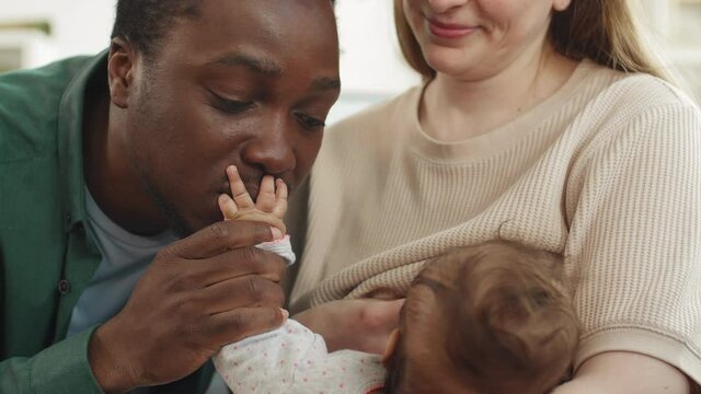 Close up of attractive Caucasian woman sitting on couch and breastfeeding her baby girl while her husband sitting nearby and kissing palm of the baby