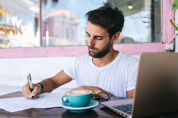 Focused man with laptop and coffee taking notes on document