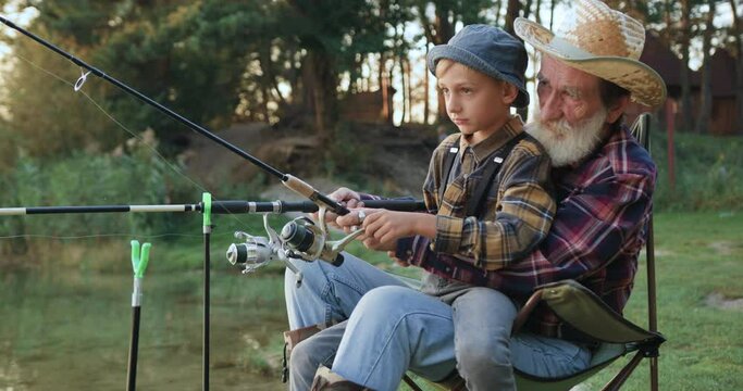 Side view of attractive confident senior bearded granddad which teaching his interested dutiful grandchild how to use rod with reel during joint fishing near the pond
