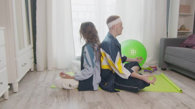 Young fit family sitting on the floor, meditating together, keeping a healthy lifestyle. Wpman uses phone while her boyfriend meditations