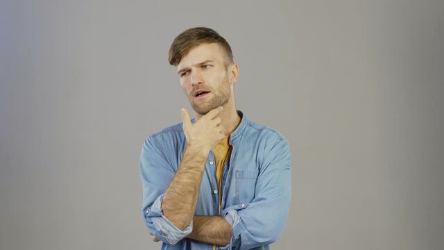 Waist up studio portrait of pensive young man thinking with his hand on chin, expressing his opinion and pointing at you against grey background