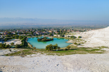 Fototapeta na wymiar Panoramic view onto lake near foot of famous travertines of Pamukkale (Turkey) & town itself. Bright green & blue color of water formed by mineral water from upper hot spring