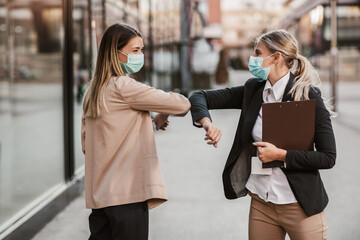 Businesswomen with safety masks greeting with elbow bump in front of office building.