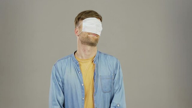 Waist up portrait of sarcastic young man looking at camera, putting on face mask and then covering eyes with it and grimacing standing against grey background
