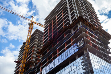 Crane is working near new building under constriction. Lower part of building is faced with mirror glass. Upper part of building is surrounded by scaffolding for workers