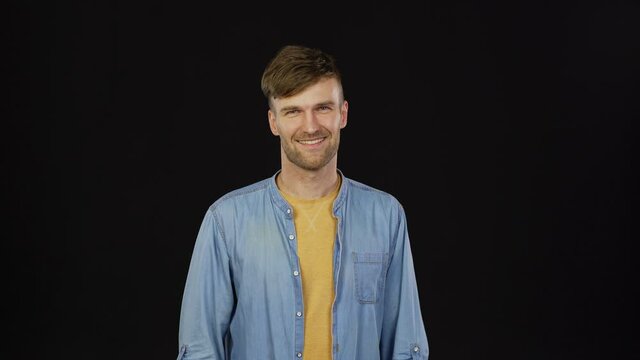 Waist up studio portrait of handsome young man turning his head, looking at camera and smiling cheerfully standing against black background