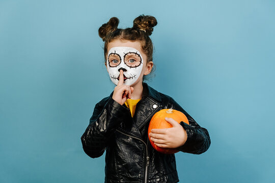 Face art and traditional Mexican holiday concept. Little girl child with orange pumpkin holding index finger at lips, asking to keep silence, dressed in Halloween costume, wears sugar skull makeup