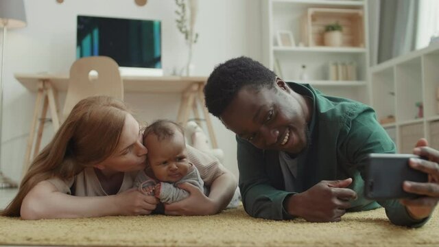 Medium shot of young Caucasian woman, African man lying on carpet in living room and making selfie with their adorable little daughter lying nearby