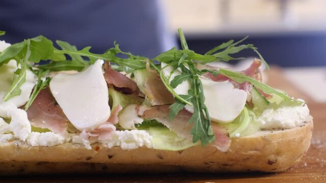 Young chef puts thin green fresh arugula lettuce leaves on a sandwich in home interior. Preparation of a morning Breakfast from a bun cucumber cheese ham. Baguette sandwich. Healthy and delicious food