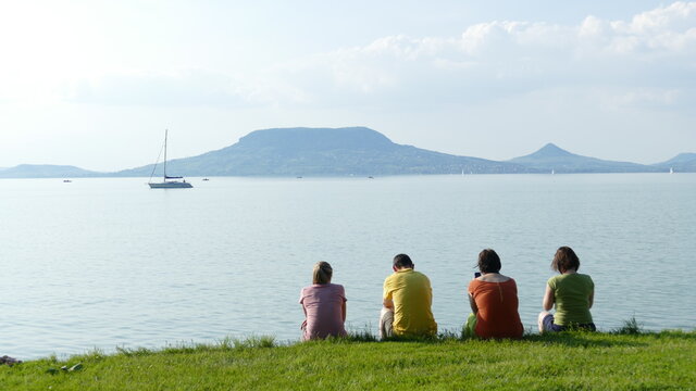 Four friends in different pastel color t-shirts sitting on the lakeside of Balaton in Hungary watching volcanic hill Badacsony and the tranquill lake in the afternoon sun
