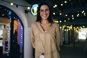 Half length portrait of cheerful female millennial in optical eyewear for eyes correction smiling at camera while standing at city urbanity, happy hipster girl in spectacles posing on leisure