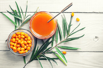 Sea buckthorn jam and fresh berries in glass jars and a green branch on a white wooden table