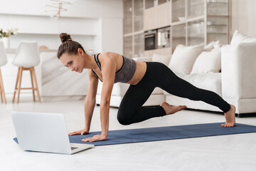 Fototapeta na wymiar Young fit girl doing fitness exercise at home while watching online tutorial lesson on laptop. Beautiful athletic girl in leggings and top practicing yoga or workout in room. Sport, healthy lifestyle