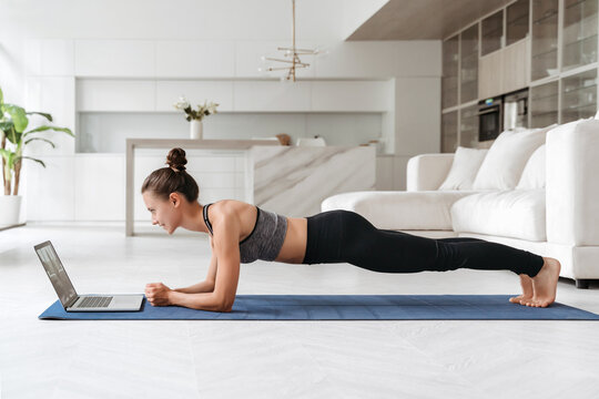 Beautiful young woman in sports uniform doing plank exercise in living room at home, watching videos on laptop computer and repeating online instructions. Online workout. Copy space. Full length side