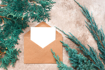 Christmas composition. Blank greeting card in a craft envelope and blue coniferous branches on a beige grunge background. Modern stationery mockup for your design. Top view, flat lay.
