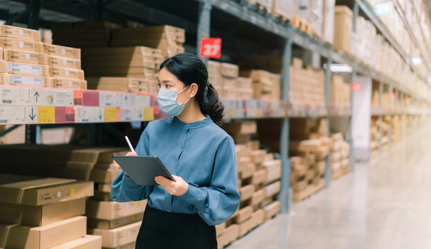 Young smart Asian business working woman wear surgical mask using digital tablet to check goods on shelves for inventory management in warehouse, Logistics business planning concept with copy space 