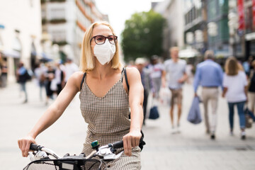 Woman riding bicycle on city street wearing medical face mask in public to prevent spreading of...