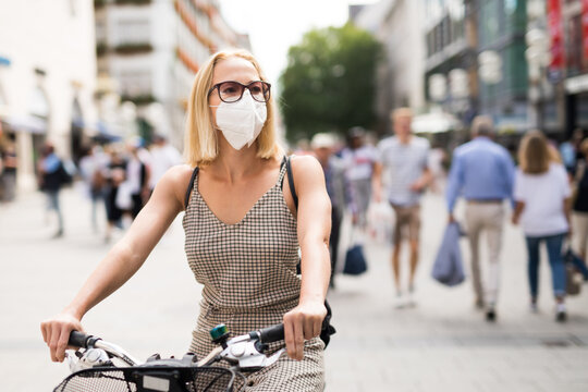 Woman riding bicycle on city street wearing medical face mask in public to prevent spreading of corona virus. New normal during covid epidemic. Social responsibility. - Powered by Adobe