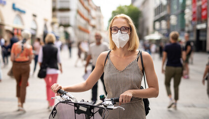 Woman walking by her bicycle on pedestrian city street wearing medical face mask in public to prevent spreading of corona virus. New normal during covid epidemic. Social responsibility. - obrazy, fototapety, plakaty